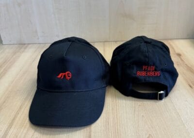 Zwei schwarze Baseballcaps mit rotem Logo und Schriftzug "Pfadi Bubenberg" auf einem hellen Holztisch, ideal für Pfadfinderfans.