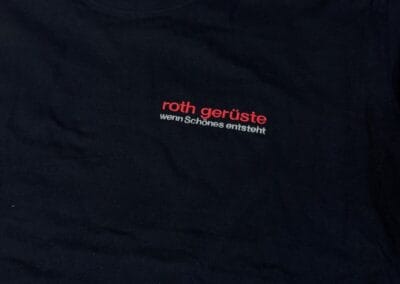 Schwarzes T-Shirt mit rotem Logo "roth gerüste" und Slogan "wenn Schönes entsteht" auf einem Holzboden. Perfekt für stilbewusste Bauprofis.