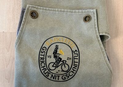 Graue Schürze mit aufgesticktem Radfahrer-Logo auf hellem Holzboden. Ideal für Grillabende und Kochabenteuer mit Freunden und Familie.