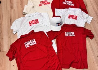 Suisse-Swimming-Kollektion: Schicke rote und weiße Hoodies und T-Shirts mit passenden Mützen auf hellem Holzboden ausgelegt. Perfekt für Fans!