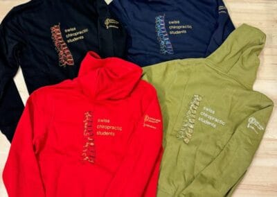 Vier bunte Kapuzenpullover mit "Swiss Chiropractic Students"-Aufdruck und Rückenmuster, die auf einem Holzboden angeordnet sind.