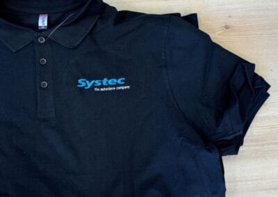 Schwarzes Polohemd mit Systec-Logo liegt auf einem Holzboden. Ideal für Arbeitskleidung oder als lässiges Büro-Outfit.