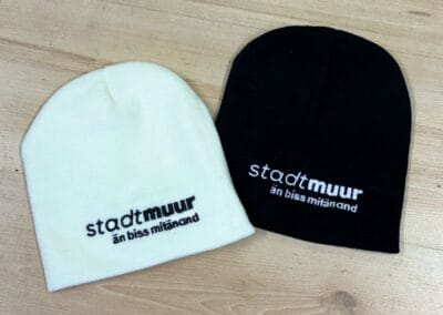 Zwei stylische Beanies, eine in Weiß und eine in Schwarz, mit dem Schriftzug "stadtmuur änn biss mitänand" auf hellem Holzboden. Perfekt für kalte Tage!